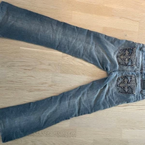 Gråblå bootcut jeans med brodyr - Snygga gråblå jeans med bootcut-modell och broderade detaljer på bakfickorna. Jeansen har klassisk femficksdesign och slitningar framtill för en cool look. Perfekta för dig som gillar en avslappnad men trendig stil. (Jag köpte dom på loppis och dom lucktar tyvärr lite funky, lite som när man går in i en källarlokal som säljer vintage och där av priser) tar absolut emot prisförslag, vill bara att någon annan får användning för dom🙏
