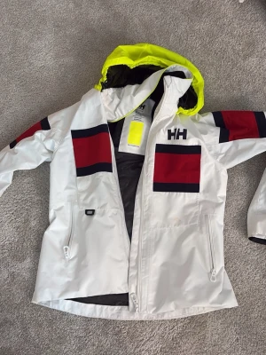 Vit Helly Hansen vindjacka med huva - Snygg vit vindjacka från Helly Hansen med röda och marinblå detaljer på bröst och ärmar. Jackan har en gul huva, dragkedja framtill och två sidofickor med dragkedja. Perfekt för blåsiga dagar och streetwear-stil.