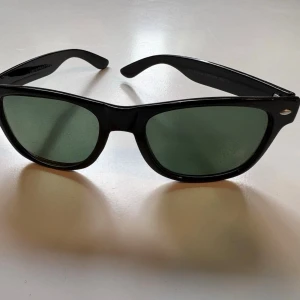 Svarta klassiska solglasögon - Snygga svarta solglasögon med gröntonade glas och tidlös wayfarer-form. Perfekta för att höja din stil på sommaren. Ramen är i plast och designen är enkel men stilren. från Henrik Stenson, Swedish design.