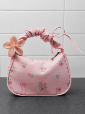 Rosa handväska med blommönster och blomklämma - Helt ny söt rosa handväska med små gröna och rosa rosor samt rosett-detaljer. Väskan har en scrunchie-inspirerad axelrem med en stor ljusrosa blomma som dekoration. Dragkedja upptill och mjuk, rundad form. Perfekt accessoar för en gullig och lekfull stil💕