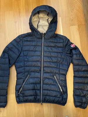 Mörkblå dunjacka från Colmar - Snygg mörkblå dunjacka från Colmar med huva och två dragkedjeförsedda fickor. Jackan har quiltad design och ett Colmar-märke på ärmen. Perfekt för kalla vinterdagar och tillverkad i lätt, värmande material.