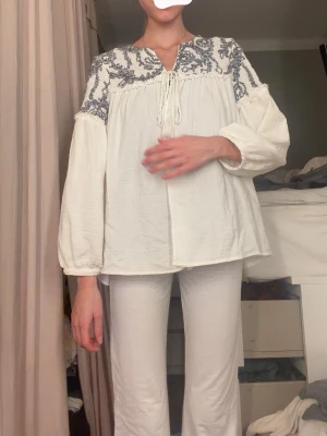 Vit blus med blå brodyr och tofsar - Luftig vit blus med blå broderade detaljer över axlar och bröst. Blusen har långa ärmar med lätt puff och knytband med tofsar vid halsen. Tillverkad i ett mjukt, vävt material. Styla för sig själv eller slänga över till stranden🥰