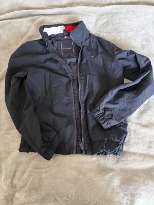 Svart vindjacka Tommy Hilfiger - Snygg svart vindjacka från Tommy Hilfiger med dragkedja framtill och elastiska muddar vid ärmar och midja. Jackan har en diskret logga på bröstet och röd detalj på insidan av kragen. Tillverkad i lätt och tåligt material, perfekt för blåsiga dagar.