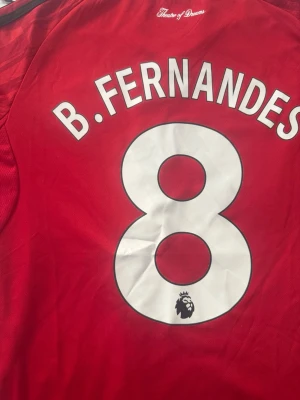 Manchester United tröja B. Fernandes 8 - Röd Manchester United matchtröja med B. Fernandes och nummer 8 på ryggen. Premier League-logga på både rygg och ärm, samt svarta och vita detaljer på ärmsluten. Tillverkad i lätt och ventilerande material, perfekt för fotbollsträning eller match.