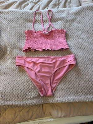 Rosa bandeau bikini med volang - Supersöt rosa bikini med bandeau-topp och smala axelband. Toppen har smockad struktur och volangkant både upptill och nedtill. Bikinitrosan är enkel och matchar toppen perfekt. Perfekt för strandhäng eller poolparty!