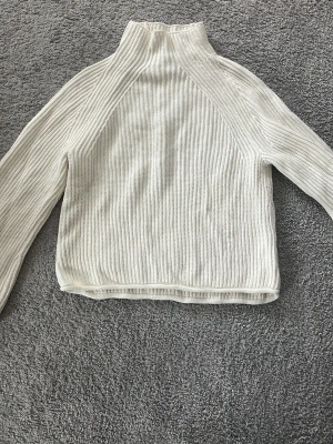 Vit ribbstickad polotröja - Säljer en stilren vit ribbstickad polotröja med hög hals och långa ärmar. Tröjan har en rak passform och är gjord i ett mjukt, tjockare material som känns skönt mot huden. Perfekt att matcha med jeans eller kjol för en clean look. Tröjan är aldrig använd och säljer eftersom jag själv inte passar i hög hals.