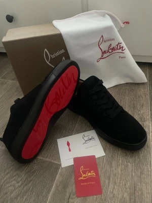 Svarta Christian Louboutin sneakers - Snygga svarta sneakers från Christian Louboutin med klassisk röd sula och diskret logga. Skorna har en låg profil, svarta skosnören och är tillverkade i mocka. Perfekta för dig som vill ha en exklusiv och stilren look.