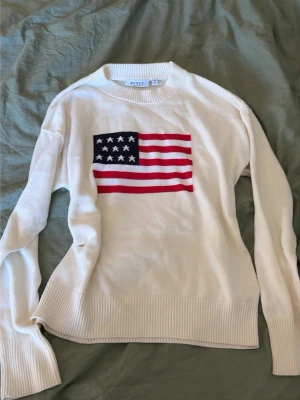 Vit stickad tröja med USA-flagga - Vit stickad tröja från RYVL med en stor amerikansk flagga i rött, vitt och blått på bröstet. Tröjan har rund halsringning, långa ärmar och breda ribbade muddar vid ärmslut och nederkant. Perfekt för en avslappnad och cool stil. Priset går att diskutera, storleken är 146-152 den är använd 1-2 ggr