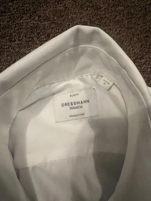 Vit slim fit skjorta från Dressmann - Klassisk vit skjorta från Dressmann Premium i slim fit-modell. Skjortan är tillverkad i ett slätt material och har en stilren design med krage och knappar framtill. Perfekt för en clean och uppklädd look.