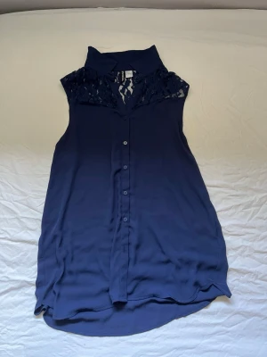 Mörkblå ärmlös blus från H&M  - Snygg mörkblå ärmlös blus från H&M med spetsdetaljer upptill och klassisk krage. Blusen har knäppning framtill och är tillverkad i ett lätt och luftigt material som känns skönt mot huden. Perfekt för en trendig och avslappnad look.