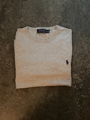 Grå tröja från Polo Ralph Lauren - Klassisk grå tröja från Polo Ralph Lauren med rund halsringning och broderad marinblå logga på bröstet. Tröjan har långa ärmar och ribbade muddar vid ärmslut och nederkant. Perfekt för en stilren och avslappnad look.