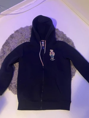 Mörkblå hoodie från New World Polo - Mörkblå hoodie med dragkedja från New World Polo Superior. Har broderad björn på bröstet och vita/röda detaljer längs dragkedjan. Klassisk huva och två fickor framtill. Perfekt för en chill och sportig stil. Använd någon gång inte mycket,  