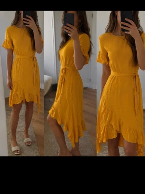 Gul omlottklänning med volang - Gul midiklänning från Soaked in Luxury i 100% viskos. Perfekt till sommarens fester eller midsommar! Omlottmodell nertill med volang och fint knytband i midjan. Korta fladdriga ärmar. Mycket fint skick.
