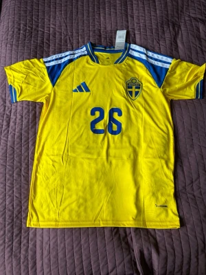 Svensk landslagströja Adidas gul/blå - Gul och blå svensk landslagströja från Adidas med nummer 26 och namnet Ali på ryggen. Tröjan har blå detaljer på axlar och ärmslut, samt det svenska fotbollsförbundets emblem på bröstet. Tillverkad i lätt funktionsmaterial (Climacool). Perfekt för fotbollsträning eller match.