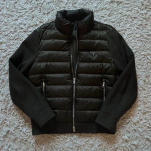 Prada | Olivgrön Cardigan  - Snygg svart pufferjacka från Prada med quiltad front och släta ärmar. Jackan har tvåvägsdragkedja, två fickor med dragkedja och diskret logga på bröstet. Materialmix med glansig front och stickade partier på ärmar och rygg. Perfekt för kalla vinterdagar. I princip oanvänd, nypris 9 tusen sek.