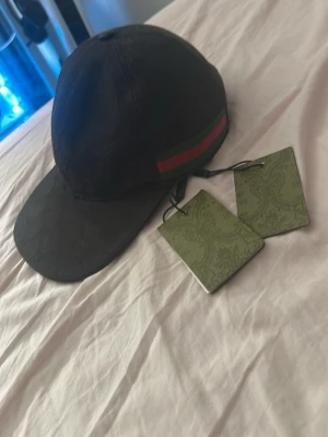 Svart keps från Gucci med röd/grön rand - Snygg svart keps från Gucci med klassisk röd och grön rand på sidan. Kepsen har böjd skärm och diskret mönster i tyget. Märkets logga finns på etiketten och kepsen har justerbar passform. Perfekt accessoar för en trendig streetwear-look.