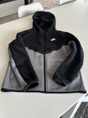 Nike Tech svart och grå XL - Snygg hoodie från Nike i svart och grå färgblock med dragkedja framtill och vit Nike-logga på bröstet. Tillverkad i mjukt material, har huva och långa ärmar. Passar perfekt för en avslappnad och sportig stil.