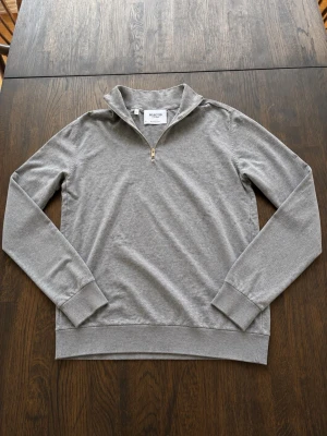 Grå halvzip tröja från Selected Homme - Snygg grå långärmad tröja från Selected Homme med halvzip och ståkrage. Tillverkad i mjukt material och har ribbade muddar vid ärmslut och nederkant. Perfekt för en clean och stilren look.