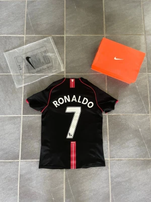 Manchester United Ronaldo fotbollströja Nike - Säljer en svart Manchester United fotbollströja från Nike med Ronaldo och nummer 7 på ryggen. Tröjan har röda detaljer, Premier League-patchar på ärmarna och AIG som sponsor på bröstet. Perfekt för dig som älskar fotboll och vill ha en ikonisk supportertröja.