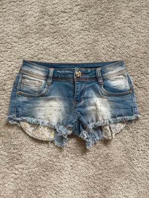 Blå jeansshorts med fransar och nitar - Säljer ett par blå lågmidjade jeansshorts med slitna fransar i nederkant och små silverfärgade nitar över hela shortsen. Shortsen har fem fickor, bälteshällor och en cool tvättad look. Insidan har ett blommigt tyg som syns lite vid benen. De är i strl xs, väldigt stretchiga. 