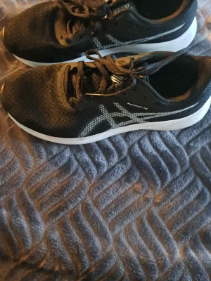 Svarta Asics sneakers med mesh - Snygga svarta sneakers från Asics med vit sula och klassiska Asics-loggan i vitt på sidan. Ovandelen är i ventilerande meshmaterial och skorna har svarta skosnören. Perfekta för dig som gillar sportig stil och vill ha bekväma skor till vardags.