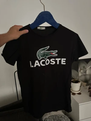 Svart Lacoste t-shirt med logga - Snygg svart t-shirt från Lacoste med den klassiska krokodilloggan och stor vit text på bröstet. T-shirten har rund halsringning och korta ärmar. Perfekt för dig som gillar stilrena och sportiga plagg med tydlig branding.