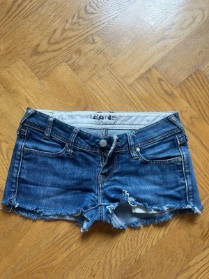 Blå jeansshorts med fransar - Säljer ett par blå jeansshorts, tror de är från Big star, med fransig, rå kant och slitningar framtill. Klassisk femficksmodell med knapp och dragkedja. Tillverkade i denim med en cool, avslappnad look. Perfekta för sommardagar och festivaler.