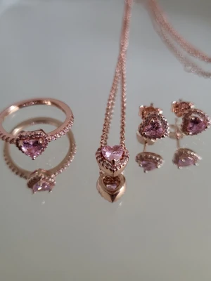 Pandora set - Snyggt Pandora smyckesset i roséguld med matchande ring(storlek 52 enligt Pandoras storleksguide), halsband och örhängen. Alla delar har hjärtformade rosa stenar omgivna av små gnistrande stenar. Perfekt för dig som gillar romantiska detaljer och vill sticka ut lite extra. En goodiebag med andra smycken och mycket mer medföljer!! 