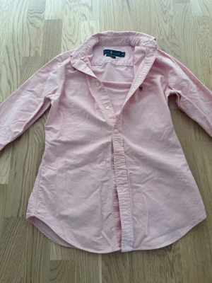 Ljusrosa skjorta från Ralph Lauren - Snygg ljusrosa skjorta från Ralph Lauren med klassisk krage och lång ärm. Skjortan har knäppning framtill och en liten broderad logga på bröstet. Tillverkad i bomull för en skön och avslappnad känsla. 