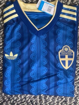 Blå Sverige fotbollströja Adidas - Säljer en blå Sverige fotbollströja från Adidas med gult märke och emblem på bröstet. Tröjan har klassiska Adidas-ränder på axlarna och ett retroinspirerat mönster. Perfekt för dig som vill stötta landslaget med stil.