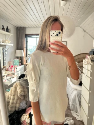 Vit stickad tröja med detaljer - Säljer en vit stickad tröja med trekvartsärmar och dekorativa knytband på ärmarna. Tröjan har en avslappnad passform och är gjord i mjukt material som känns skönt mot huden. Perfekt för dig som gillar stilrena plagg med en twist.