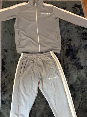 Grått tracksuit från Palm Angels - Säljer ett grått tracksuit-set från Palm Angels med vita detaljer. Setet består av en zip-jacka med hög krage och två fickor samt matchande byxor med resår i midjan och vita ränder längs sidorna. Båda delarna har Palm Angels-loggan tryckt i vitt.