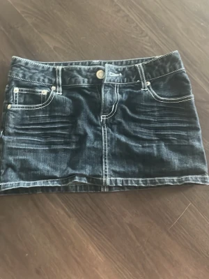 Mörkblå jeanskjol - Snygg mörkblå jeanskjol i kort modell med tydliga vita kontrastsömmar. Kjolen har klassiska fem fickor, knapp och dragkedja framtill samt dekorativa sömmar på bakfickorna. Helt oanvänd