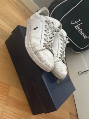 Vita Polo Ralph Lauren sneakers - Snygga vita sneakers från Polo Ralph Lauren med svart detalj på hälen och klassisk logga på sidan. Skorna har vita snören med Polo-tryck och är tillverkade i läder. Perfekta för en clean och stilren look.  Vid köp så får dom en bra och nogran rengöring inann som skickad och de kommer göra att dom ser mycket nyare ut