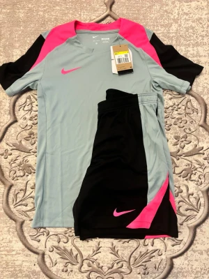Nike träningsset ljusblå/svart/rosa - Snyggt träningsset från Nike med t-shirt och shorts. T-shirten är ljusblå med svarta och rosa detaljer, och har en rosa Nike-logga på bröstet. Shortsen är svarta med ljusblå och rosa inslag samt rosa Nike-logga. Perfekt för fotboll eller gym. Tillverkad i lätt och ventilerande material. Shorts är L och tröjan är S, passar bra till någon som vill ha lite större byxor 