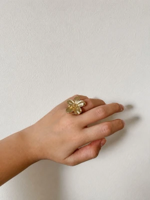Stor guldfärgad blomring - Unik ring i guldfärgad metall med en stor, detaljerad blomma som motiv. Blomman har fem blad med fina räfflade detaljer som ger en cool och iögonfallande look. Defekt: lite färg är borta, ojämn justerbar cirkel. Sitter fint ändå!
