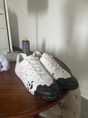  Maison Margiela svart paint splatter - Maison Margiela Sneakers – Storlek 43  Säljer ett par Maison Margiela sneakers i storlek 43. Vit bas med svart, handmålad/splash-inspirerad design som ger skorna en unik och exklusiv look.  Skorna är i nyskick och endast använda en gång.  * Märke: Maison Margiela * Storlek: 43 * Färg: Vit/Svart * Skick: Nyskick – använda en gång dust bag medföljer Samnt kartong