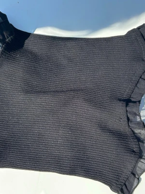 Svart ribbad bandeau topp med snörning - Svart ribbad topp med korta ärmar och snygga volangdetaljer vid ärmslut och hals. Toppen har snörning i sidan som ger en cool och trendig look. Perfekt för dig som gillar stilrena men ändå unika plagg.