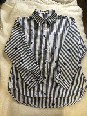 Randig skjorta med blå hjärtan - Snygg skjorta i vitt och blått med vertikala ränder och små blå broderade hjärtan. Klassisk krage, knäppning framtill och lång ärm. Jätte bra om man vill vara lite extra fin eller bara ha något fint till varadags. Helt ny men utan prislapp, passade inte mig tyvärr