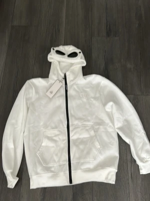 Vit hoodie med glasögon från C.P. Company - Säljer en vit hoodie från C.P. Company med hel dragkedja och ikoniska inbyggda glasögon i huvan. Hoodien har känguruficka framtill och ribbade muddar. Perfekt för dig som vill sticka ut med en unik streetwear-look.