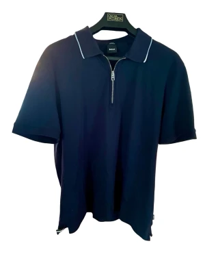 Marinblå kortärmad t-shirt från BOSS - Snygg marinblå t-shirt från BOSS med korta ärmar och vit piping på kragen. Tröjan har en klassisk krage och en half zip-dragkedja framtill. Tillverkad i mjuk bomull för en bekväm känsla och stilren look. Plagget är i nyckick använd 2-3 gånger, passar tyvärr inte mig. Inga fläckar eller slitage förekommer. Liten i storleken ”true size” är snarare L än XL. 