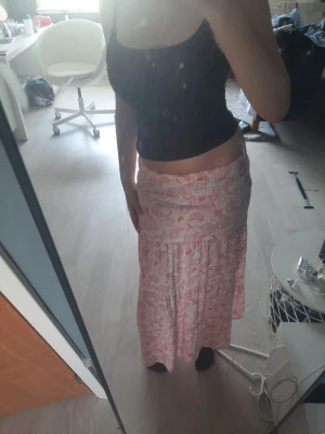 Lång paisleymönstrad kjol i rosa - Säljer en lång kjol med paisleymönster i rosa och vitt. Kjolen har resår i midjan och flera volangpartier som ger en luftig och bohemisk känsla. Perfekt för dig som gillar en avslappnad men ändå snygg stil.