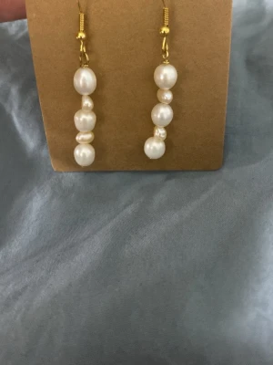 Guldiga örhängen med vita pärlor - Säljer ett par eleganta örhängen med guldfärgade krokar och vita pärlor i olika storlekar på rad. Örhängena har en klassisk och stilren look som passar till många outfits. Perfekta för dig som gillar en tidlös accessoar med pärldetaljer.