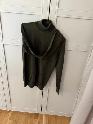 Olivgrön polotröja i stickat material från J.Lindberg - En stilren olivgrön stickad polotröja med lång ärm och ribbade muddar vid ärmslut och nederkant. Tröjan har en klassisk polokrage och är tillverkad i ett mjukt, tunt material som passar perfekt till lager-på-lager-stil. Använts några enstaka tillfällen.