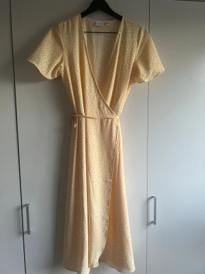 Ljusgul omlottklänning från Vila - Söt lång omlottklänning från Vila i ljusgul färg med vitt prickigt mönster. Klänningen har korta ärmar, knyt i midjan och v-ringad hals. Perfekt för varma dagar och har en luftig, flowy känsla. 💕💕