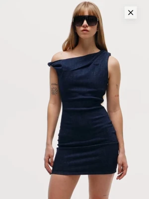 Mörkblå oneshoulder jeansklänning - Trendig mörkblå kort jeansklänning med oneshoulder-design och figurnära passform. Klänningen har en asymmetrisk axel och är tillverkad i ett stretchigt denimtyg som ger en cool och modern look. Den är i storlek S från Bershka. Helt ny med prislapp