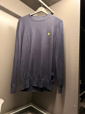 Stickad Lyle & Scott - En stilren mörkblå tröja med rund hals och lång ärm. Tröjan har en liten gul broderad logga på bröstet och ribbade muddar vid ärmslut och nederkant. Perfekt för dig som gillar enkel och klassisk stil.