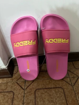 Rosa slides med gul text - Säljer ett par rosa slides med bred rem och gul text på ovansidan. Sandalerna har en enkel och sportig design, perfekt för avslappnade dagar. Tillverkade i syntetmaterial med mjuk innersula för extra komfort.
