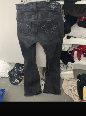 Svarta True Religion jeans bootcut - Säljer ett par svarta jeans från True Religion med bootcut-modell och ikoniska sömmar på bakfickorna. Jeansen har klassisk femficksdesign och metallknappar. Perfekta för dig som gillar streetwear och vill sticka ut lite extra.