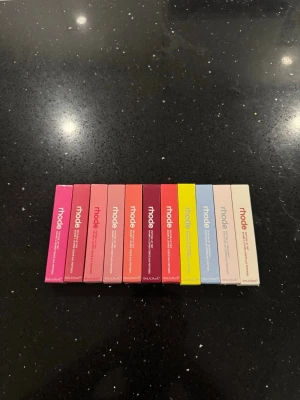 Rhode Peptide Lip Treatment set 12 st - Säljer ett set med Rhode Peptide Lip Treatment i flera olika smaker och färger, bland annat Watermelon Slice, Cinnamon Roll, Sugarberry, Passionfruit Jelly, Guava Spritz, Raspberry Jelly, Peach Pit, Jelly Bean, Ribbon, Sugar Cookie, Shortcake och Rhode Vanilla. Förpackningarna är i olika färger och har en modern, stilren design.